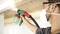 Una persona sta levigando un soffitto in legno con un utensile elettrico. Indossa occhiali protettivi e una maschera antipolvere.