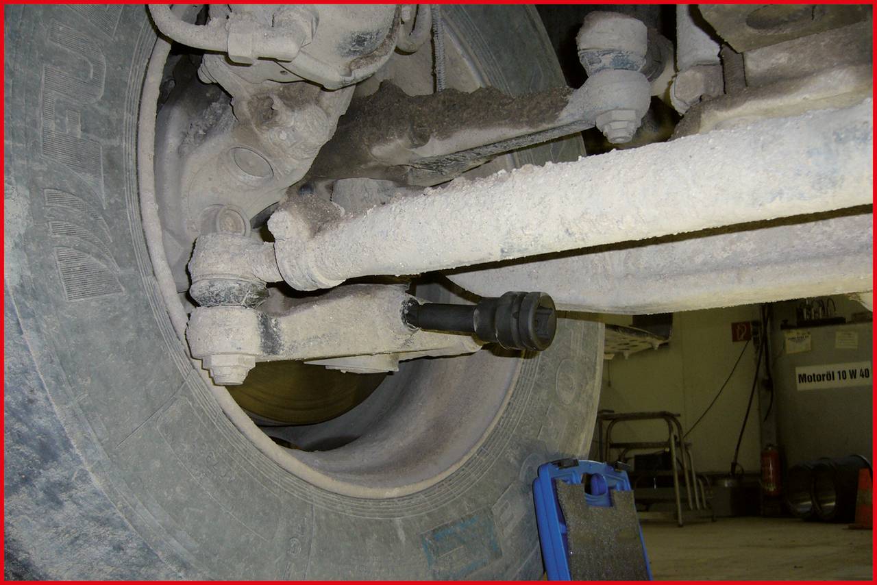Primo piano di un pneumatico di un camion e della sua sospensione, ricoperti da uno strato di polvere. Sullo sfondo sono visibili attrezzature di officina.