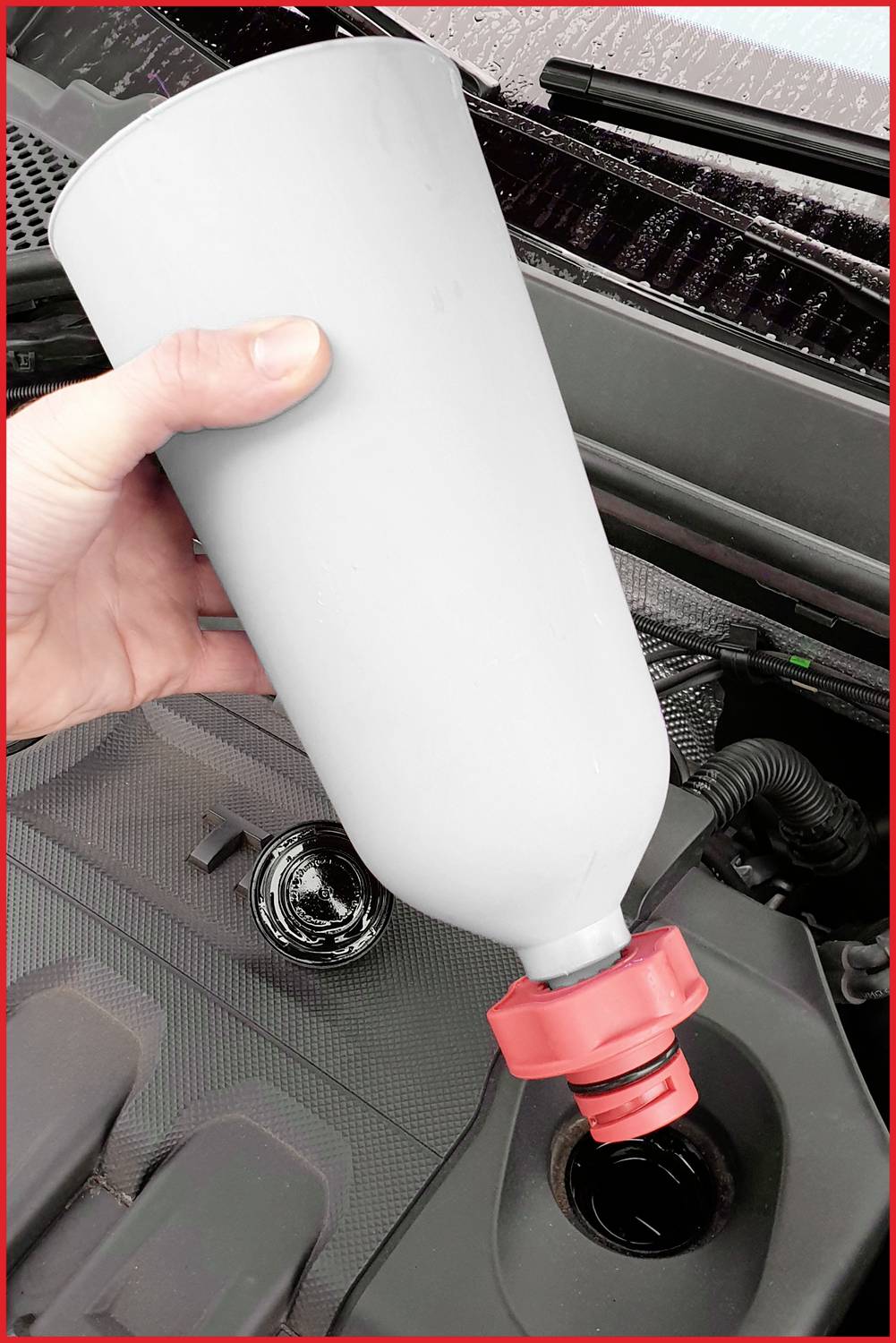 Una mano versa liquido da una grande bottiglia grigia con tappo rosso nel foro di riempimento del motore del veicolo.
