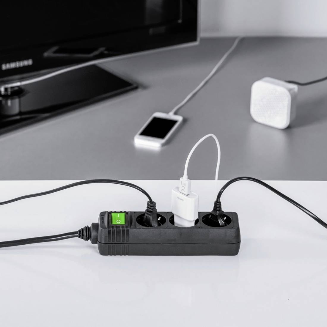 Una presa multipla con diversi dispositivi collegati, tra cui un caricabatterie per cellulare e un interruttore illuminato, su un tavolo. Sullo sfondo uno smartphone e un altoparlante.