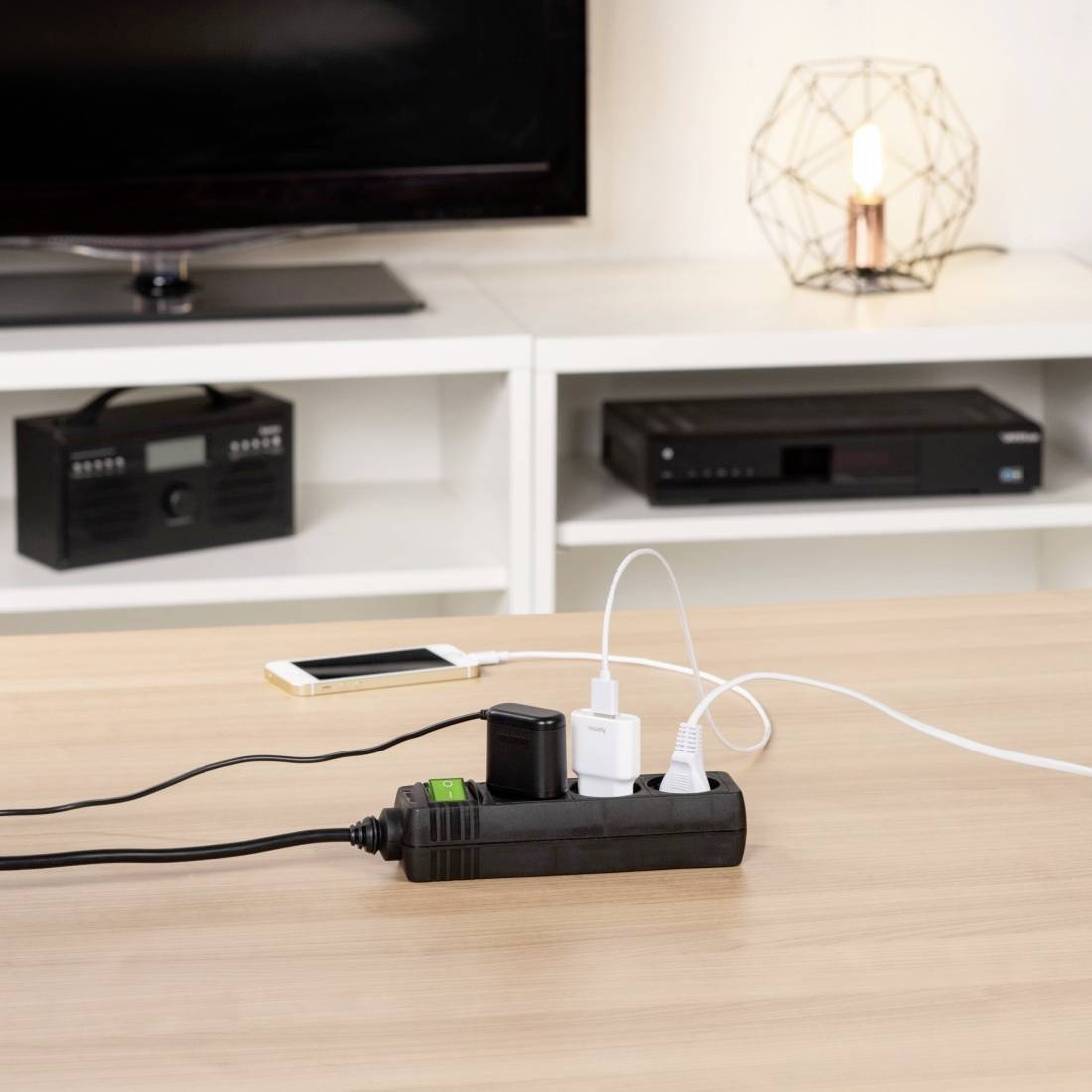Presa multipla su un tavolo con quattro caricabatterie collegati per smartphone e altri dispositivi elettronici, con una lampada sullo sfondo.