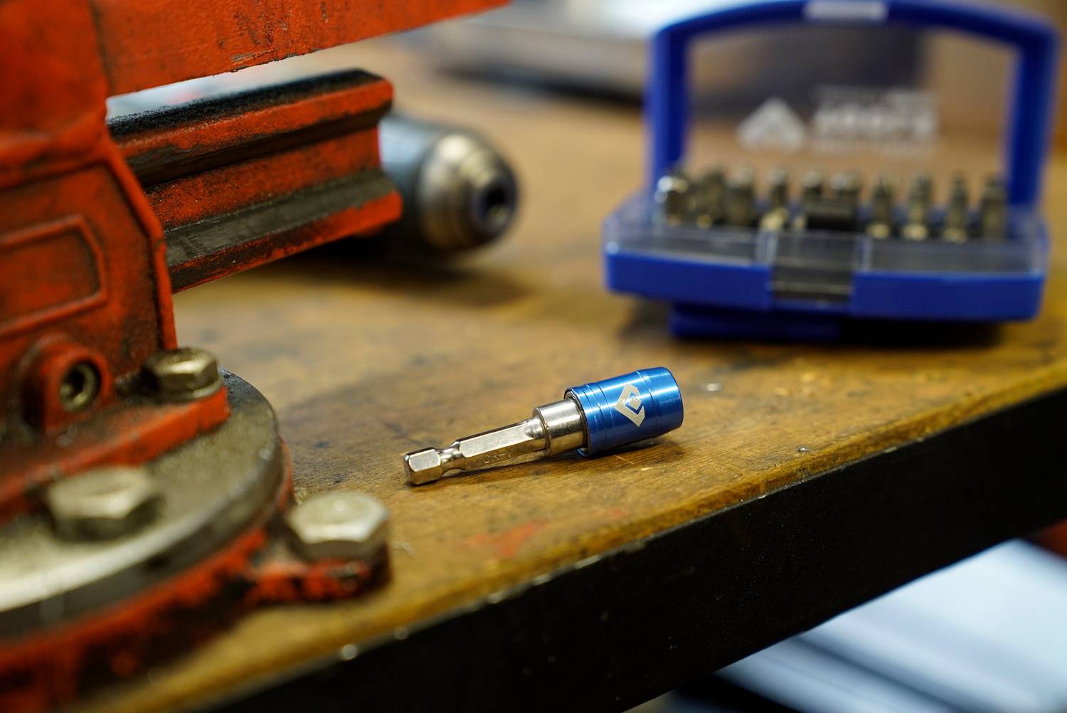 Un bit per cacciavite blu è posizionato su un banco da lavoro, circondato da una morsa e una scatola per bit sullo sfondo.