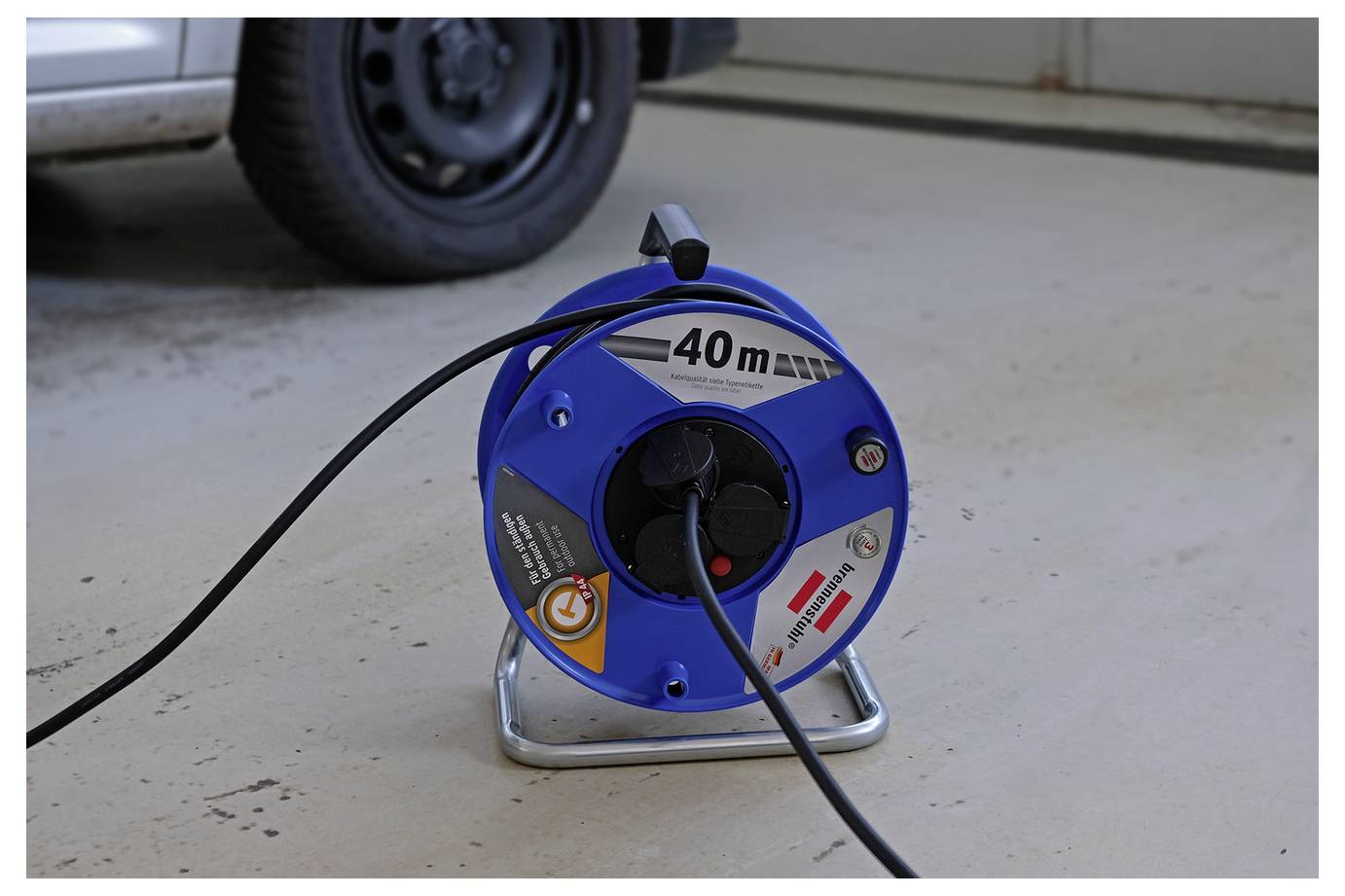 Avvolgicavo blu con una prolunga nera su un pavimento di garage, etichettato "40 m", vicino a uno pneumatico di un veicolo.