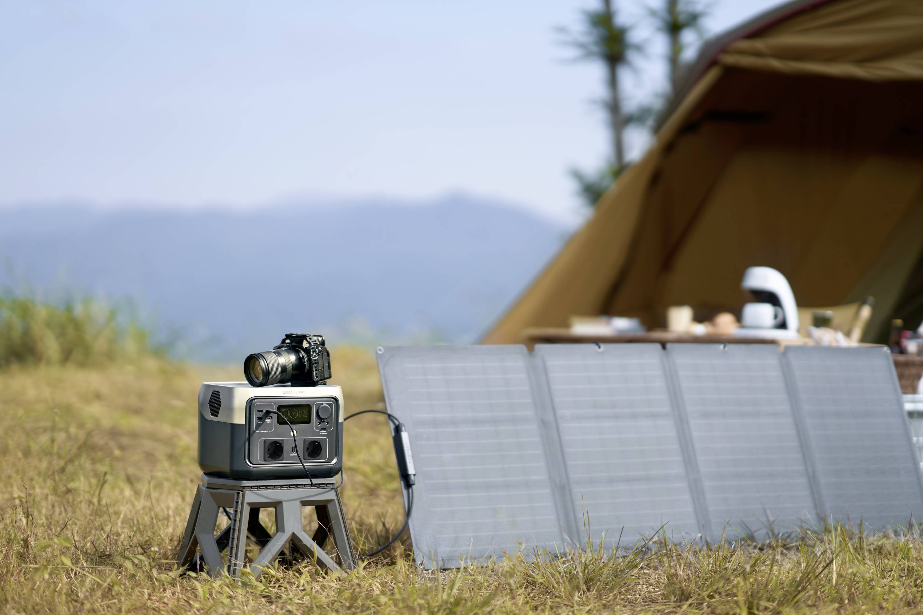 Un pannello solare accanto a una tenda da campeggio, con una telecamera e un caricabatterie collegati. Sullo sfondo si vedono delle montagne.