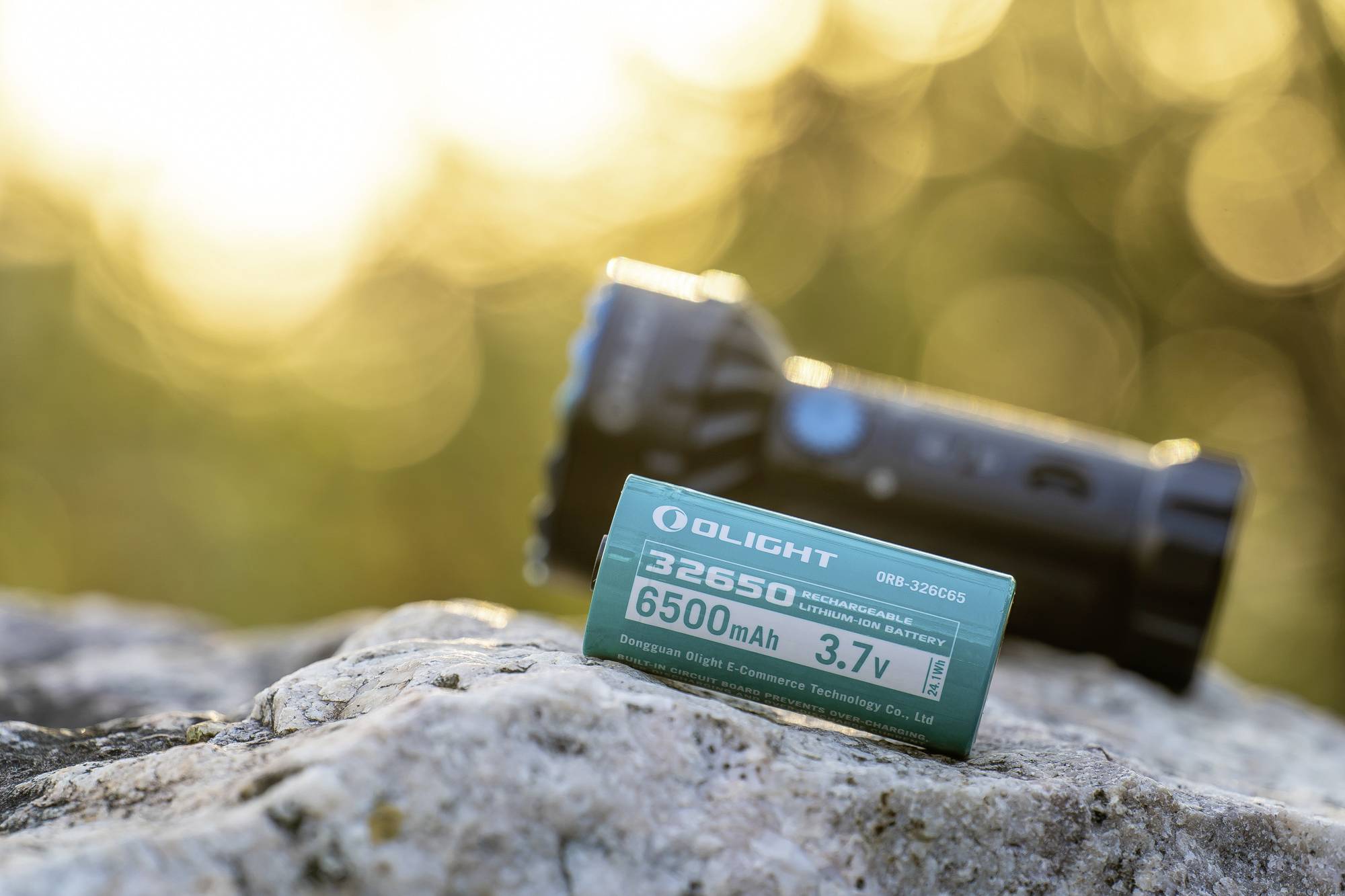 Una batteria 32650 da 6500 mAh e 3,7 V poggia su una pietra, sullo sfondo sfocato una torcia e riflessi dorati di luce.