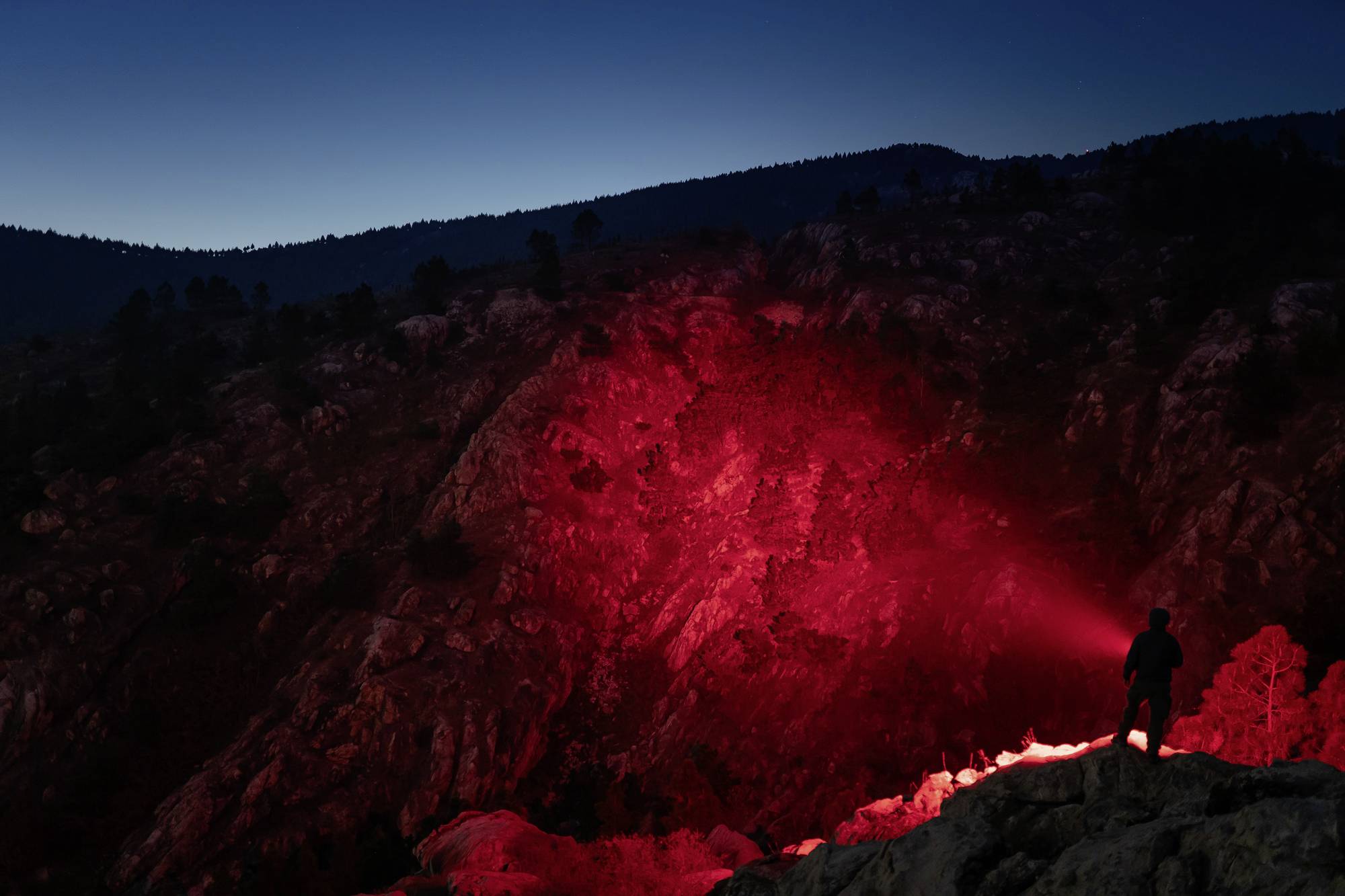 Una persona è in piedi su una collina rocciosa e illumina le rocce con una torcia rossa. Il cielo è al crepuscolo.