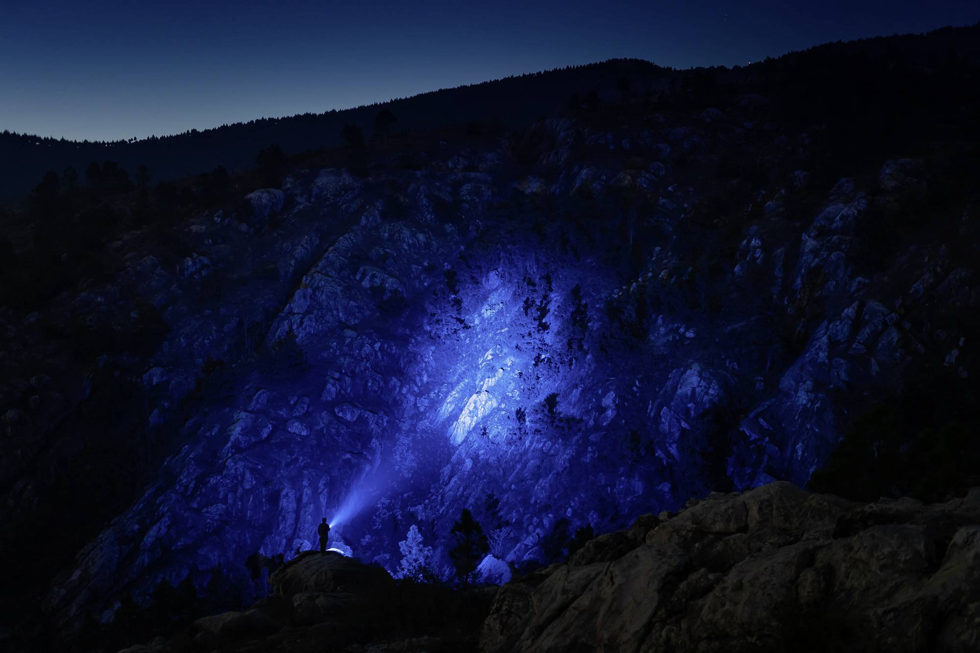 Una persona è in piedi su un masso e illumina con una torcia elettrica una parete rocciosa di montagna nell'oscurità, creando un cono di luce blu.