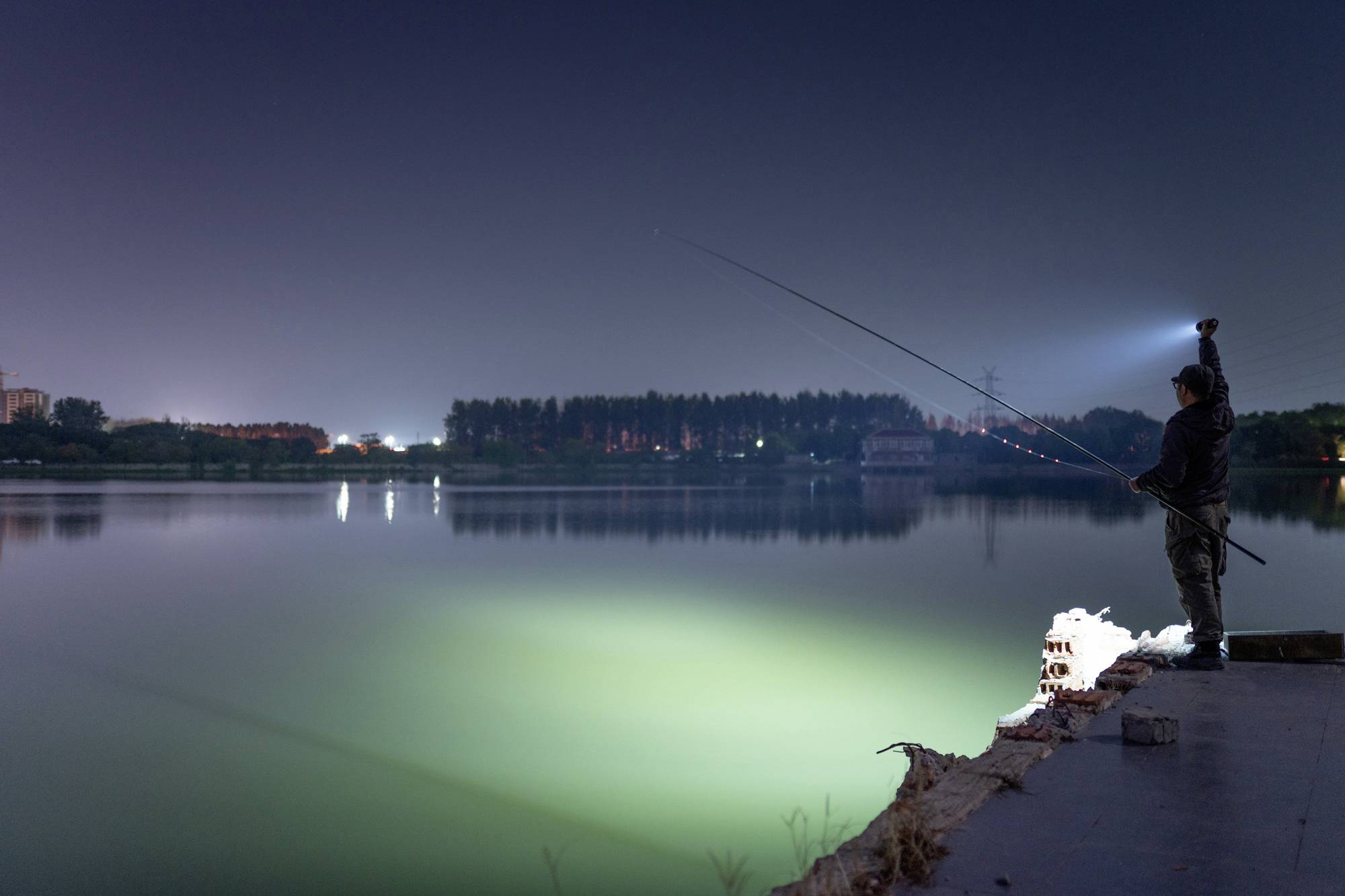 Una persona pesca di notte su un lago tranquillo, illuminata da una lampada; acqua riflettente e alberi sullo sfondo.