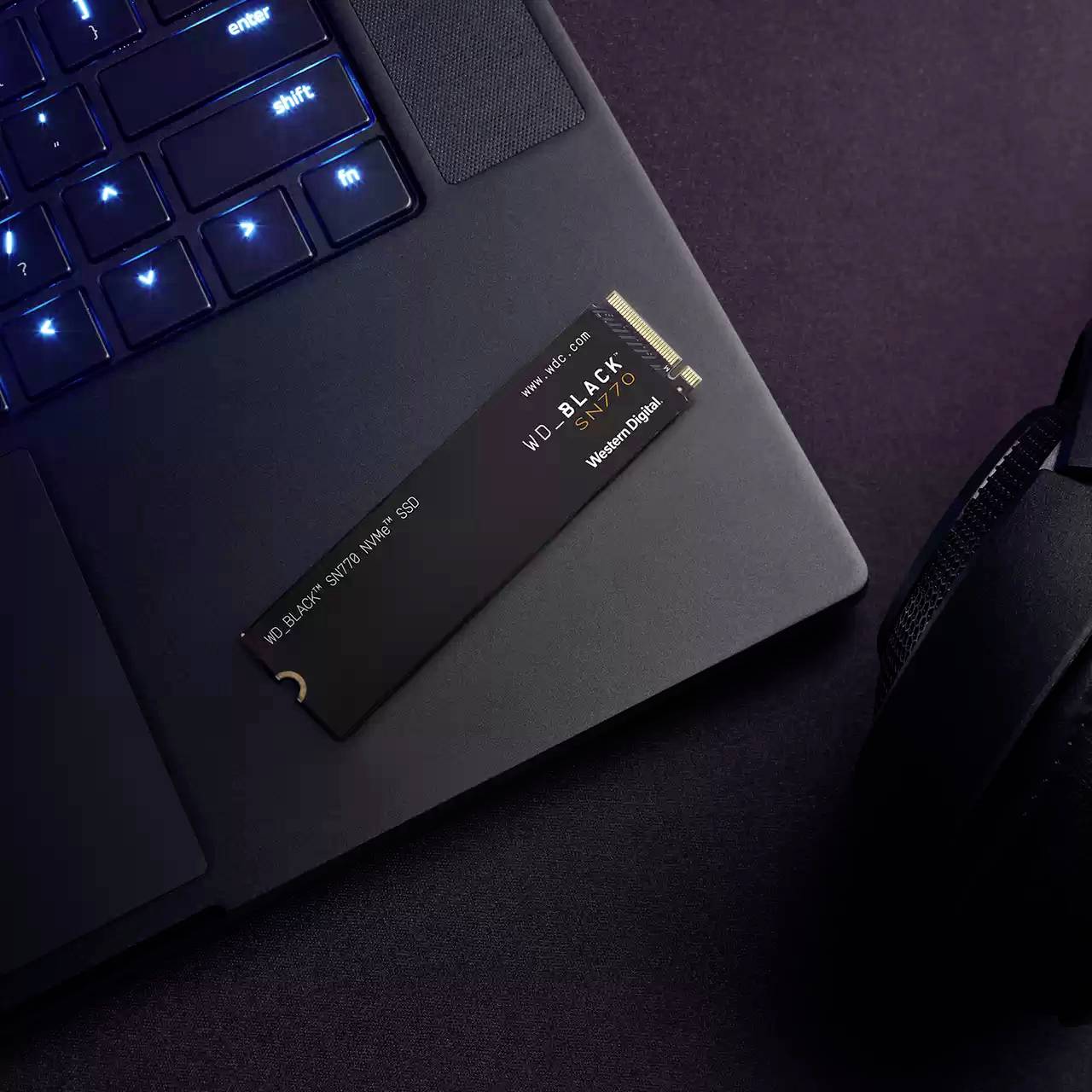 Un laptop con tastiera retroilluminata è posizionato su un tavolo. Un hard disk esterno è poggiato sulla tastiera. Accanto si trovano delle cuffie.