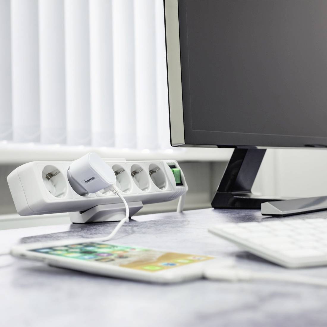 Un tavolo da lavoro con un monitor acceso, una ciabatta elettrica con un caricabatterie inserito e uno smartphone in primo piano.