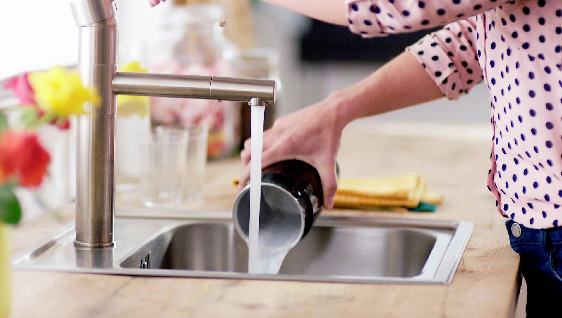 Una persona sta sciacquando un bicchiere sotto l'acqua corrente in un lavandino della cucina. Sullo sfondo ci sono dei fiori e un asciugamano da cucina giallo.