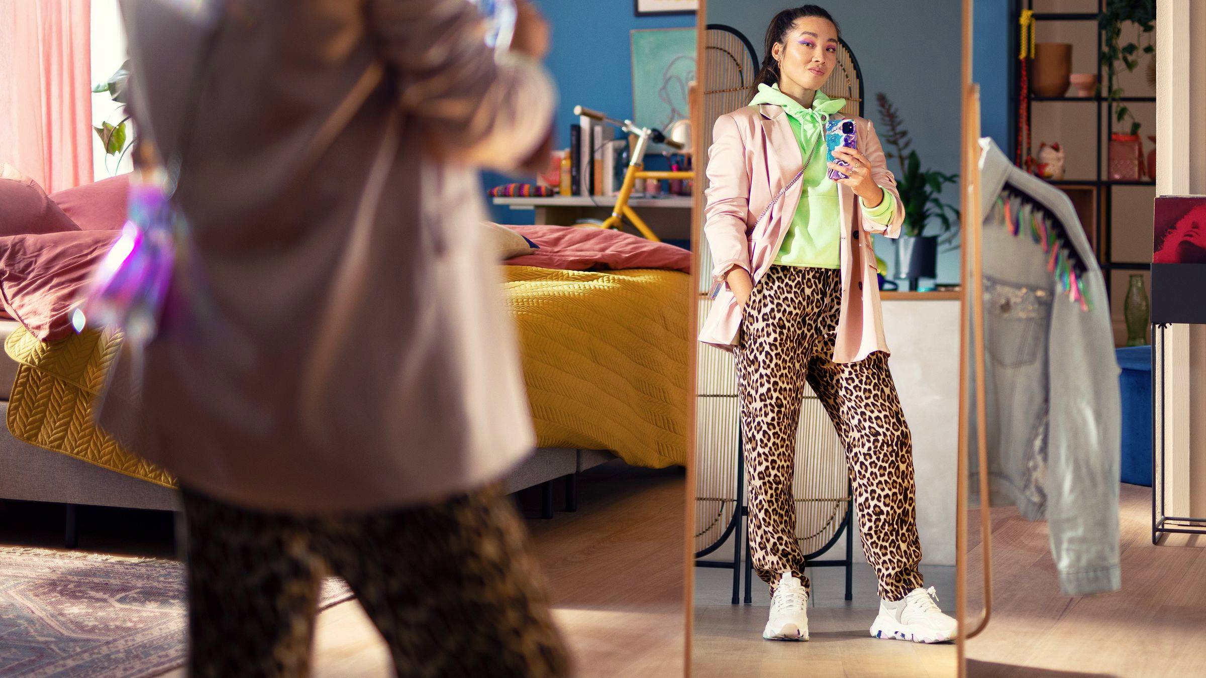 Una persona è davanti a uno specchio, indossa un blazer rosa, un felpa verde e pantaloni leopardati. Tiene in mano un cellulare per un selfie. Camera da letto sullo sfondo.