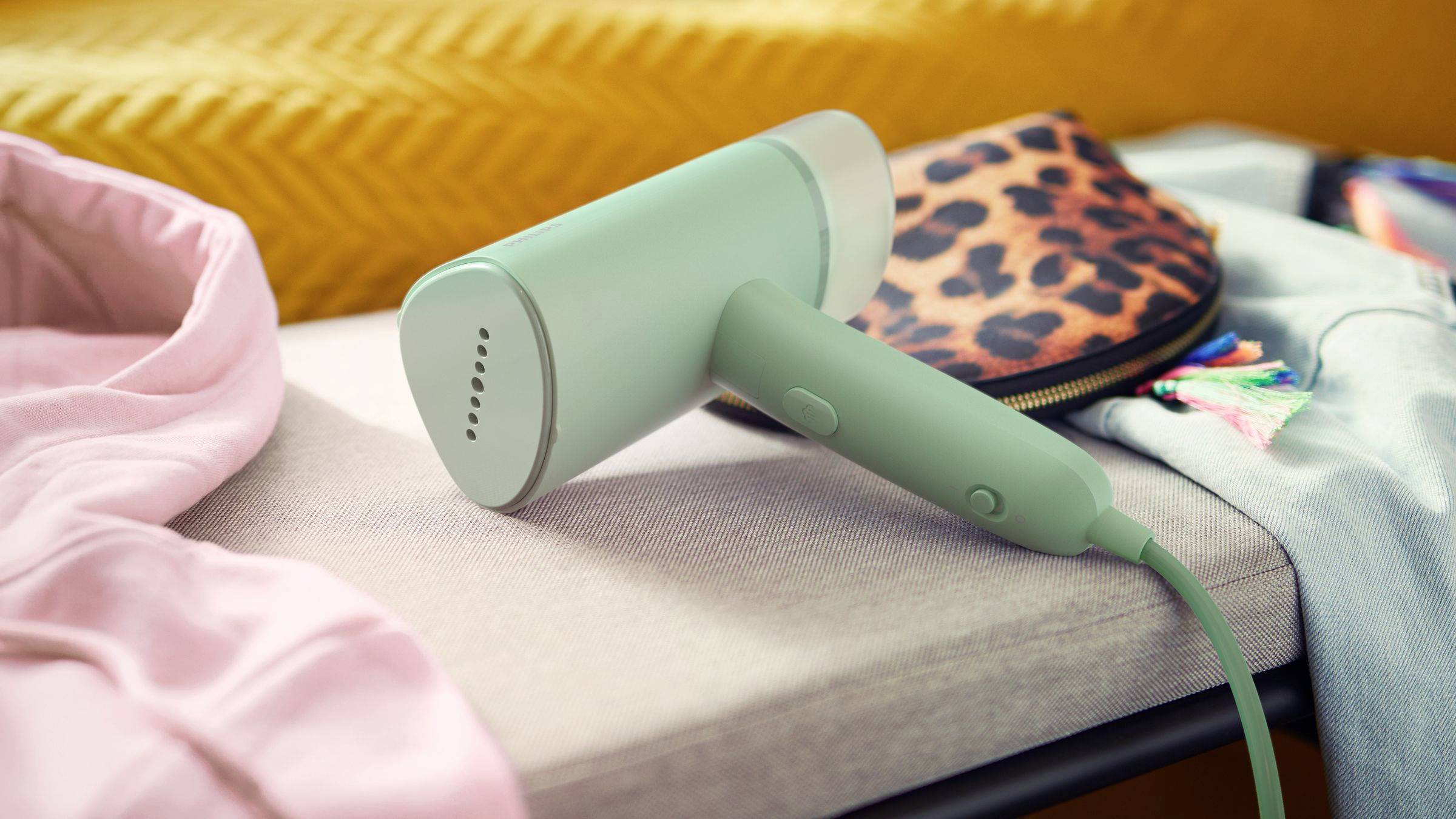 Un ferro da stiro a vapore verde è poggiato su una superficie di tessuto tra capi di abbigliamento colorati. Sullo sfondo si intravede una borsa con stampa leopardata.
