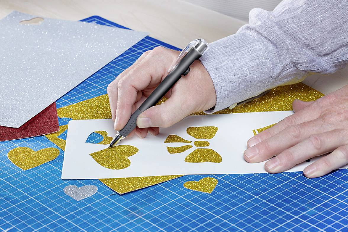 Una persona taglia forme dorate da carta glitterata con un bisturi su un tappetino da taglio. Le forme sono cuori e fiocchi.