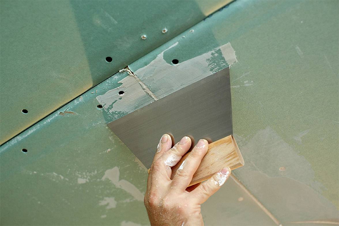 Una mano stucca le giunture di una parete in cartongesso verde con una spatola. Sono visibili tre viti.
