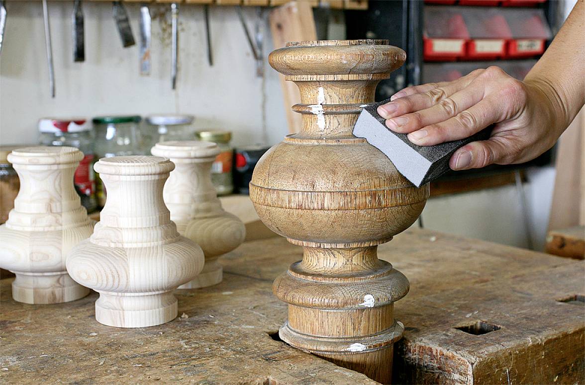 Una persona sta levigando un pezzo di legno tornito in un laboratorio. Sullo sfondo si vedono altri pezzi di legno e attrezzi.