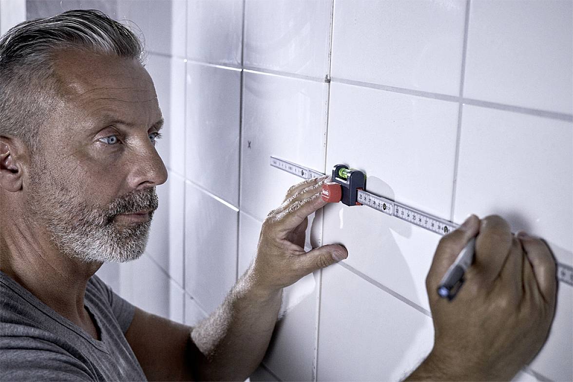 Un uomo con la barba misura le piastrelle su un muro utilizzando un telemetro laser e un righello.