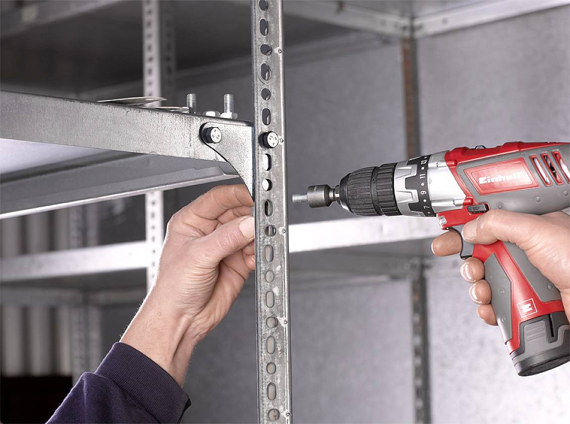 Una persona avvita un bullone in uno scaffale metallico utilizzando un avvitatore a batteria rosso e nero per stabilizzare la struttura.