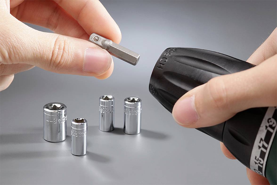 Mano che tiene un avvitatore a batteria e una punta; quattro chiavi a tubo sono posizionate accanto. Viene mostrato il cambio degli utensili.