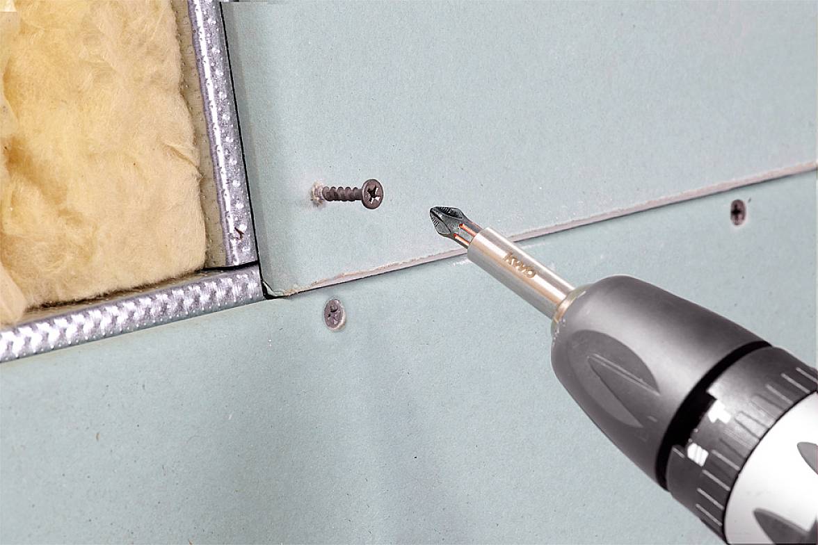 Un trapano avvitatore sta inserendo una vite in una lastra di cartongesso fissata a una cornice metallica. Dietro si vede del materiale isolante.
