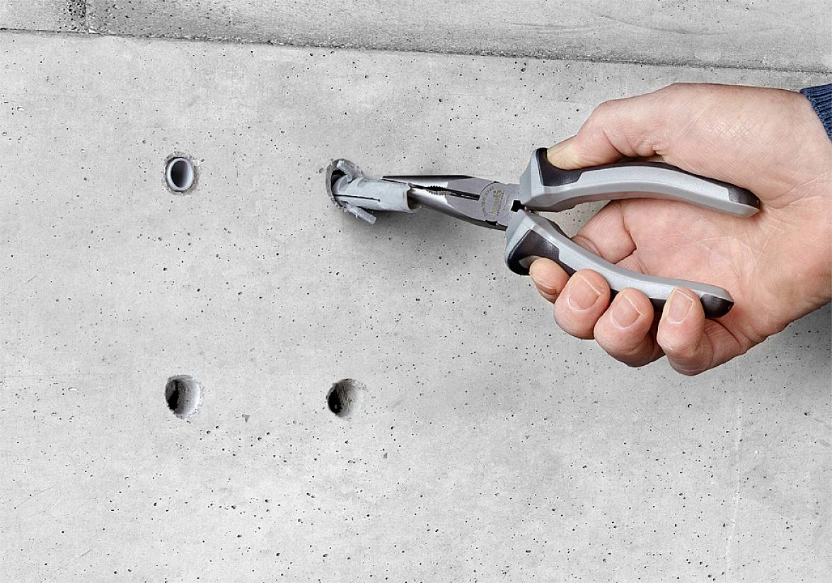 Una mano tiene una pinza e estrae un tassello da un muro in cemento. Sono visibili altri tre fori nella parete.