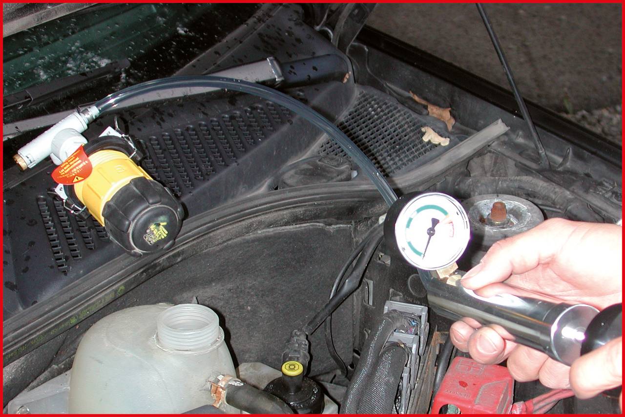 Una mano tiene un dispositivo per test di pressione nel vano motore di un'auto. Una valvola gialla è fissata a un tubo.