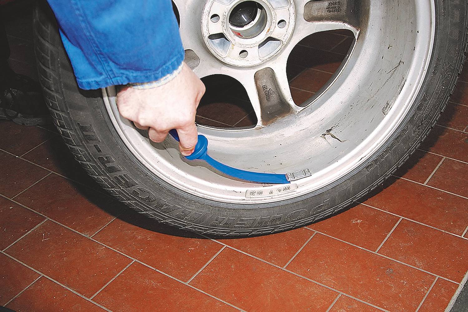 Una persona misura la profondità del battistrada di un pneumatico dell'auto con un utensile blu, poggiato su un pavimento di piastrelle rosse.