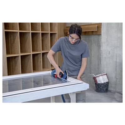Una persona utilizza un attrezzo elettrico per levigare i bordi di una cornice di legno su un tavolo in un laboratorio. Scaffalature e un secchio sono sullo sfondo.