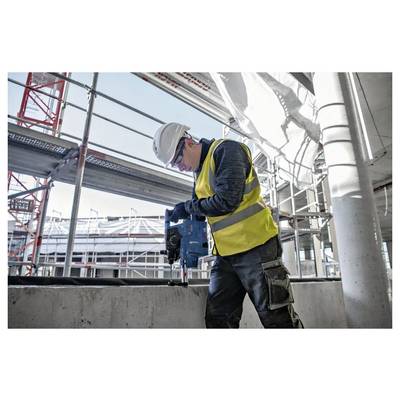 Un lavoratore edile con un giubbotto di sicurezza giallo e un elmetto aziona un trapano su una superficie in calcestruzzo in un cantiere con ponteggi.