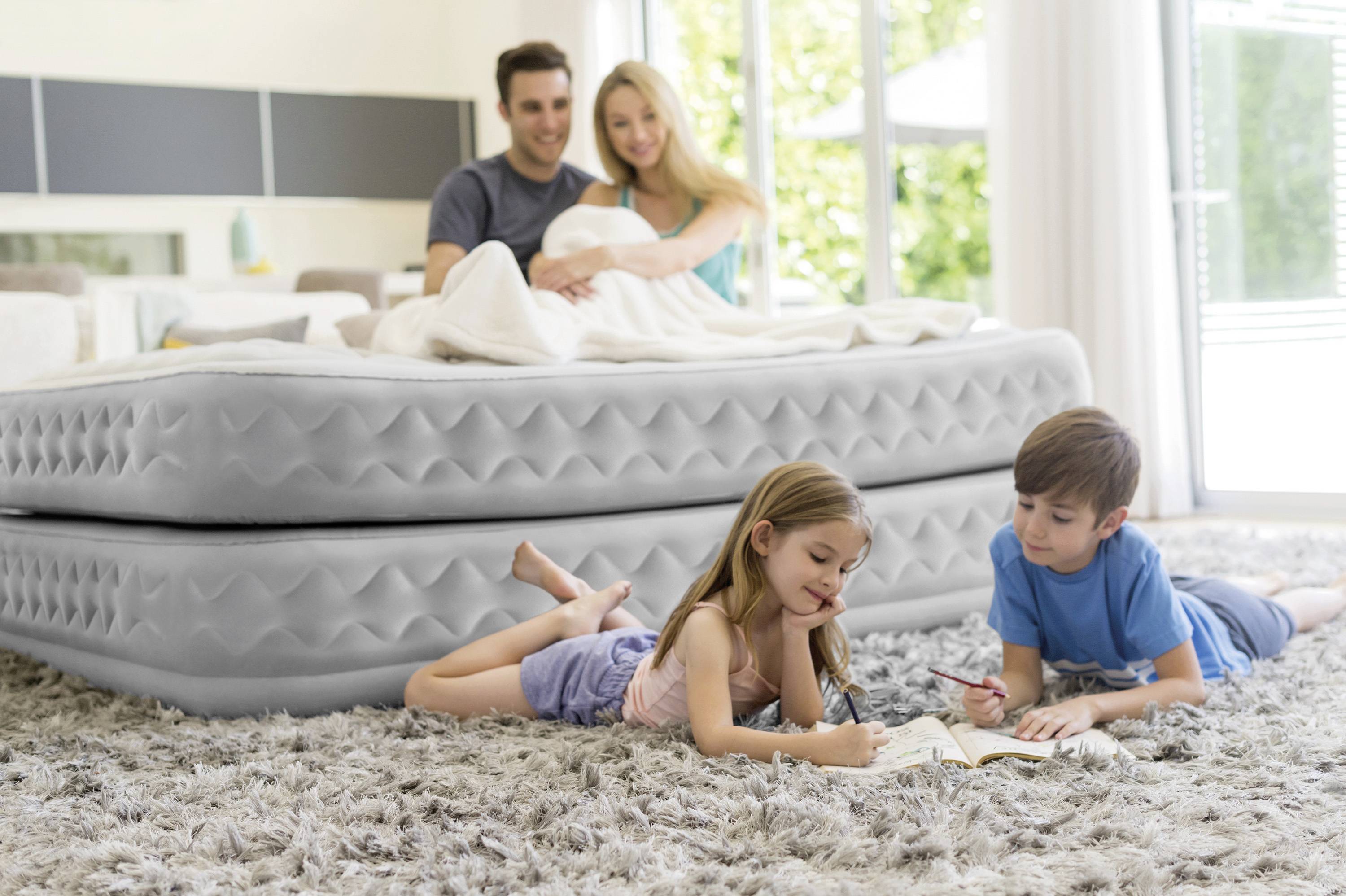 Una famiglia in soggiorno; i genitori sono seduti su un grande materasso ad aria, mentre due bambini sono sdraiati sul tappeto e stanno scrivendo in un libro.