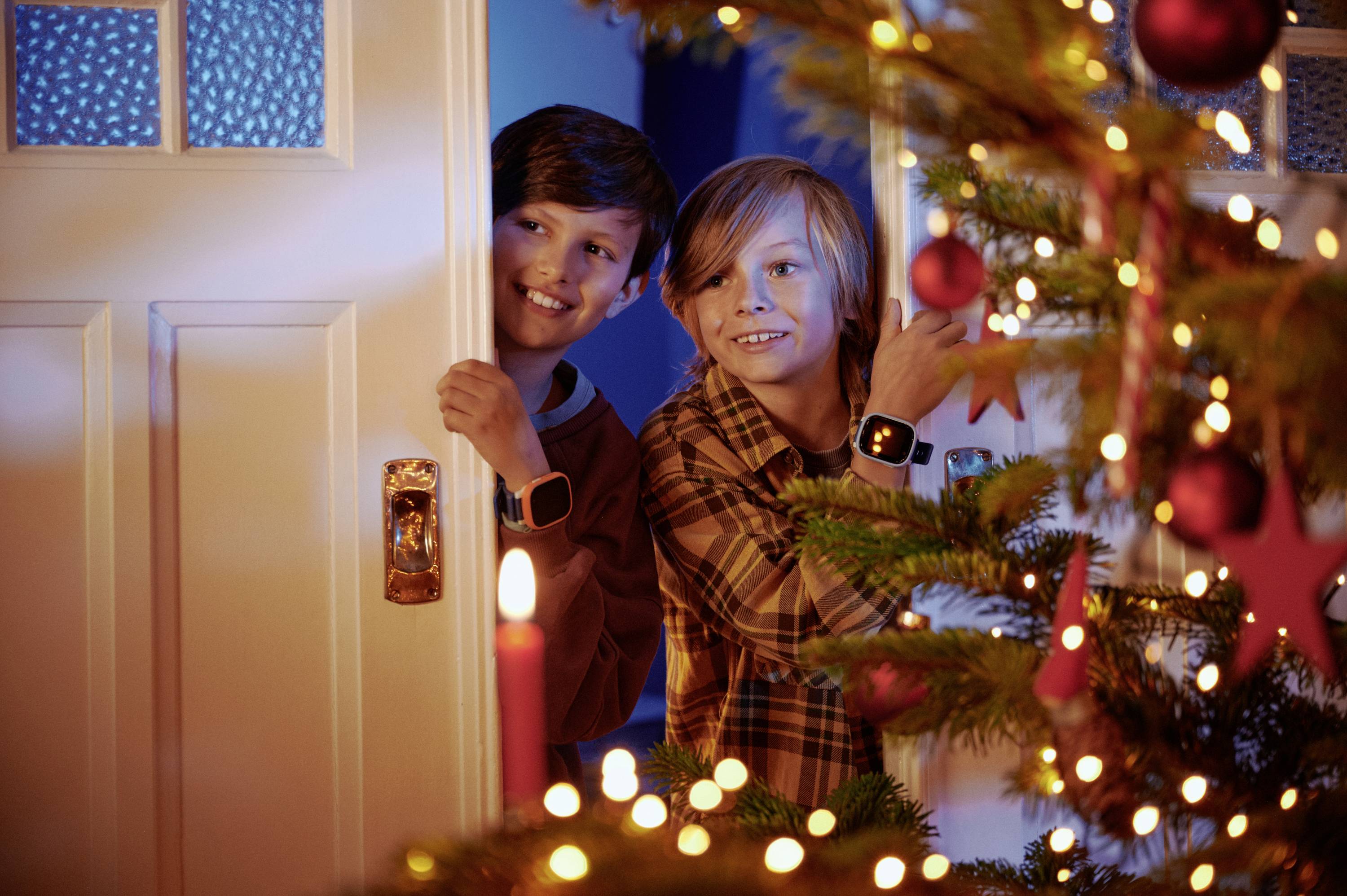 Due bambini spuntano ridendo da dietro una porta che conduce a una stanza addobbata per Natale con un albero illuminato.