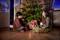 Un ragazzo e una ragazza siedono sorridenti davanti a un albero di Natale addobbato con regali sotto. Un'atmosfera festosa riempie la stanza.
