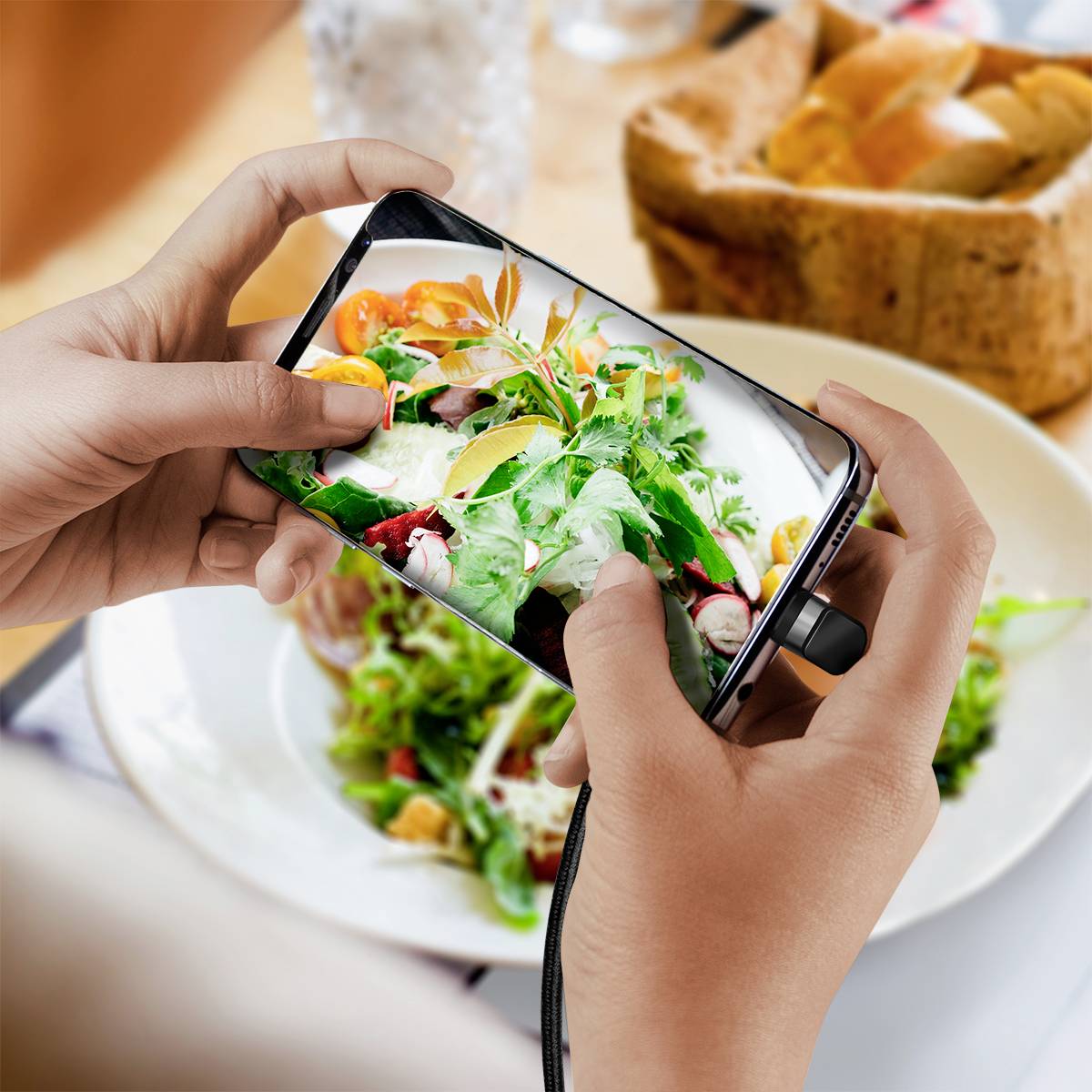 Una persona tiene uno smartphone e fotografa un piatto di insalata colorato su un piatto. Sullo sfondo si vedono un bicchiere e del pane.