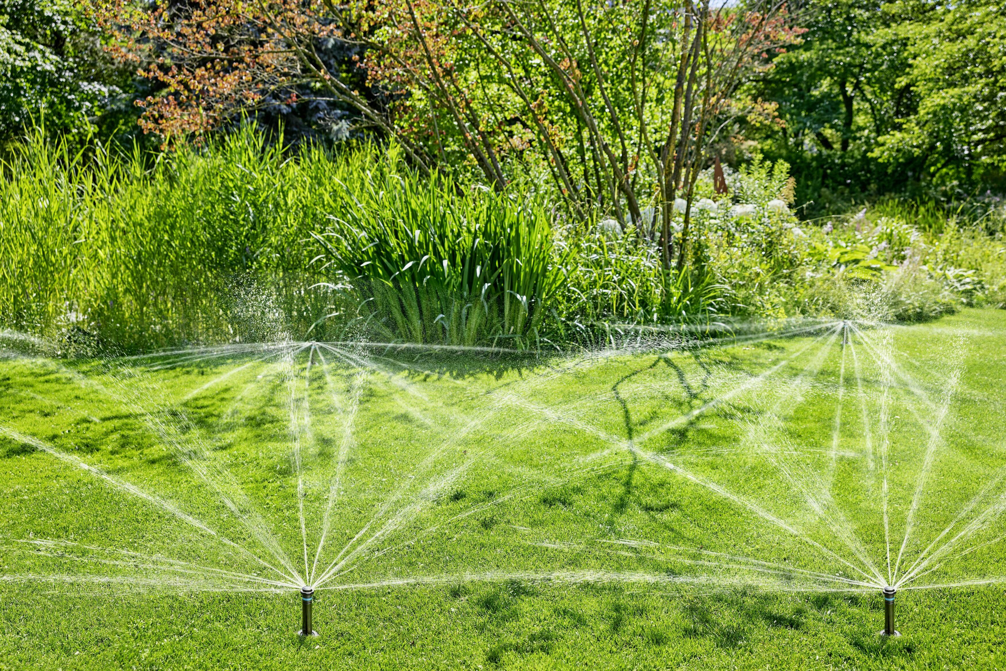 Un giardino con erba verde viene irrigato da due spruzzatori in movimento, circondato da alberi e cespugli sullo sfondo.