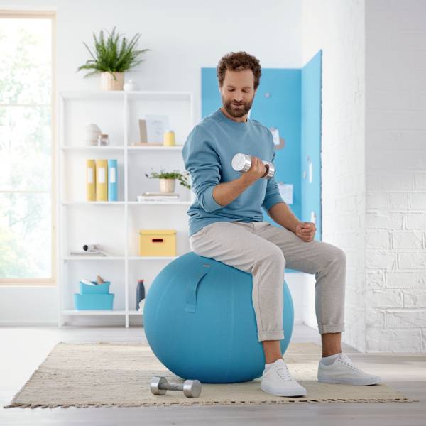 Un uomo è seduto su un pallone da ginnastica in un salotto mentre solleva un manubrio. Sullo sfondo si trovano una libreria e una finestra.