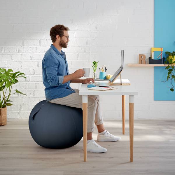 Un uomo è seduto su un pallone da ginnastica accanto a una scrivania, lavora su un laptop. Ufficio moderno e luminoso con piante.
