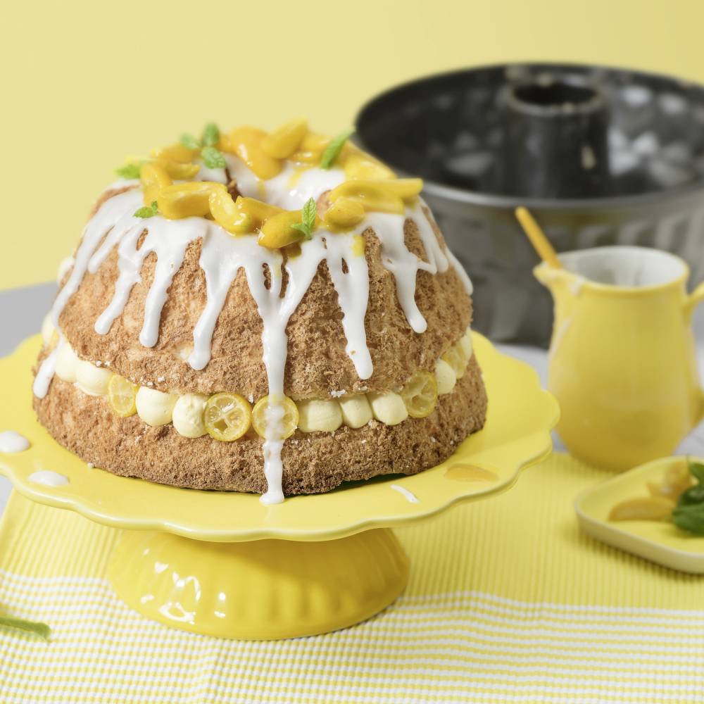 Una torta su un supporto giallo, decorata con glassa e fette di limone; sullo sfondo una brocca e una teglia da forno.