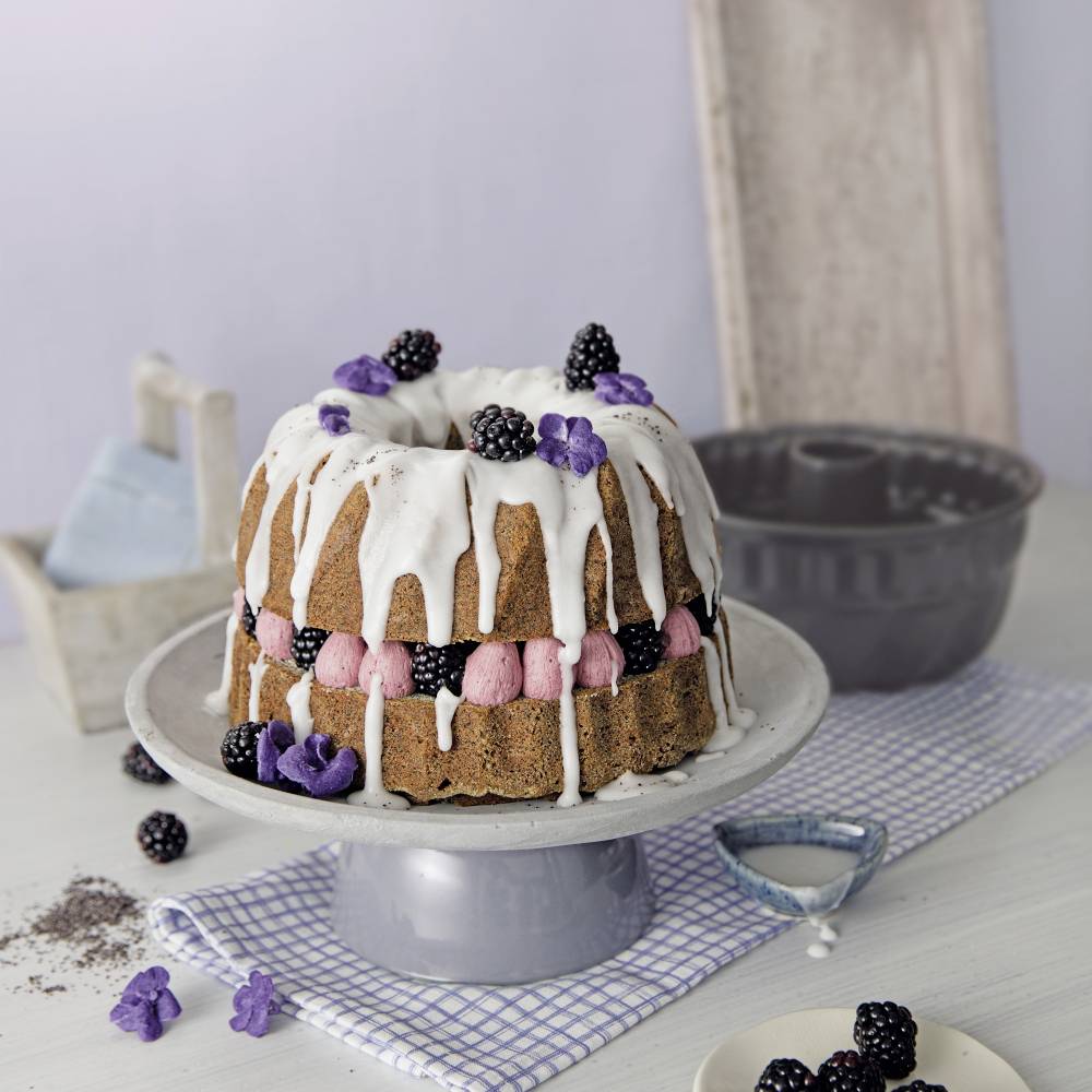 Un dolce Bundt decorativo con glassa bianca, decorato con more e fiori viola, è poggiato su un tavolo. Sullo sfondo è visibile uno stampo per ciambella.
