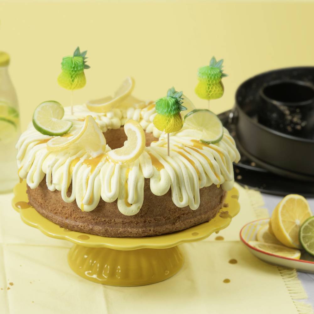 Torta con glassa e fette di limone, decorata con bastoncini di ananas, su un piatto giallo; sullo sfondo limonata.