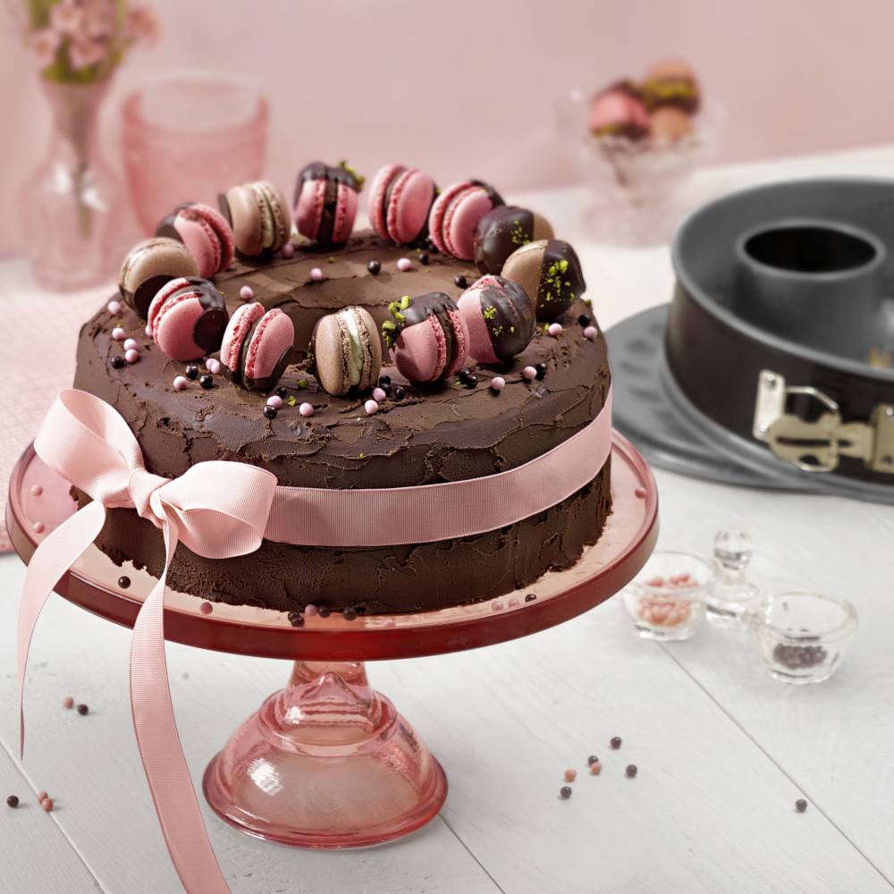Una torta al cioccolato con un nastro rosa su un supporto rosa, decorata con macarons. Sullo sfondo una tortiera e spezie.