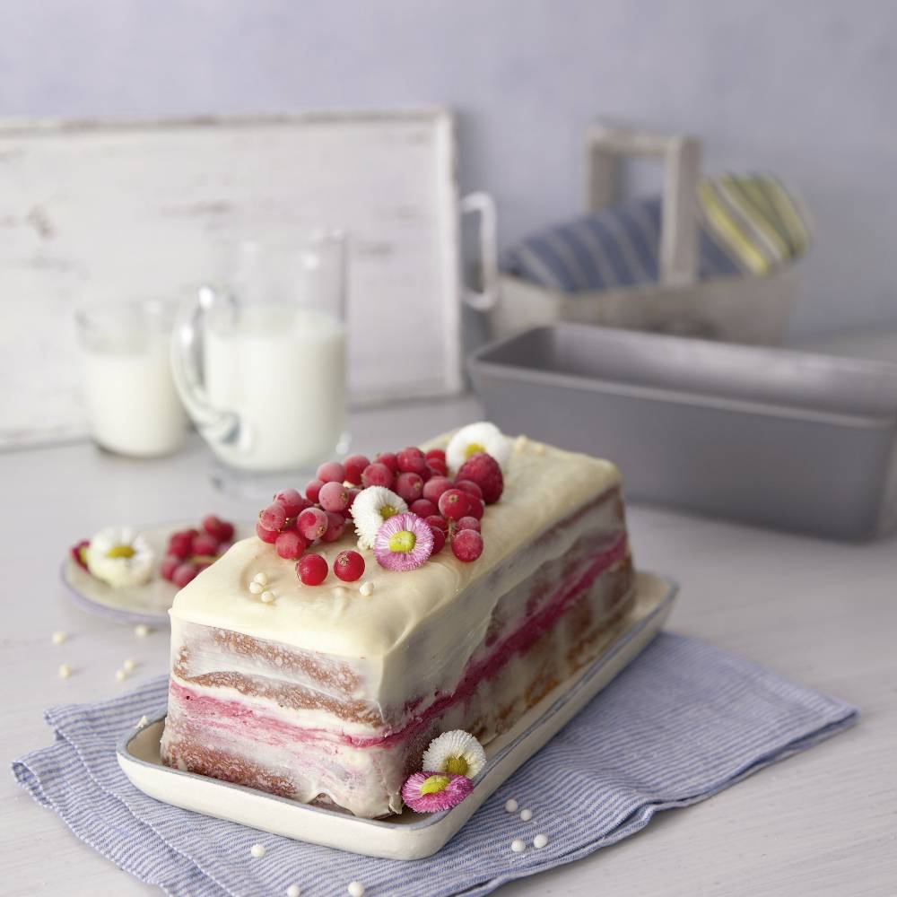 Una torta con glassa bianca e ribes rossi in cima, poggia su un tavolo con un bicchiere di latte sullo sfondo.