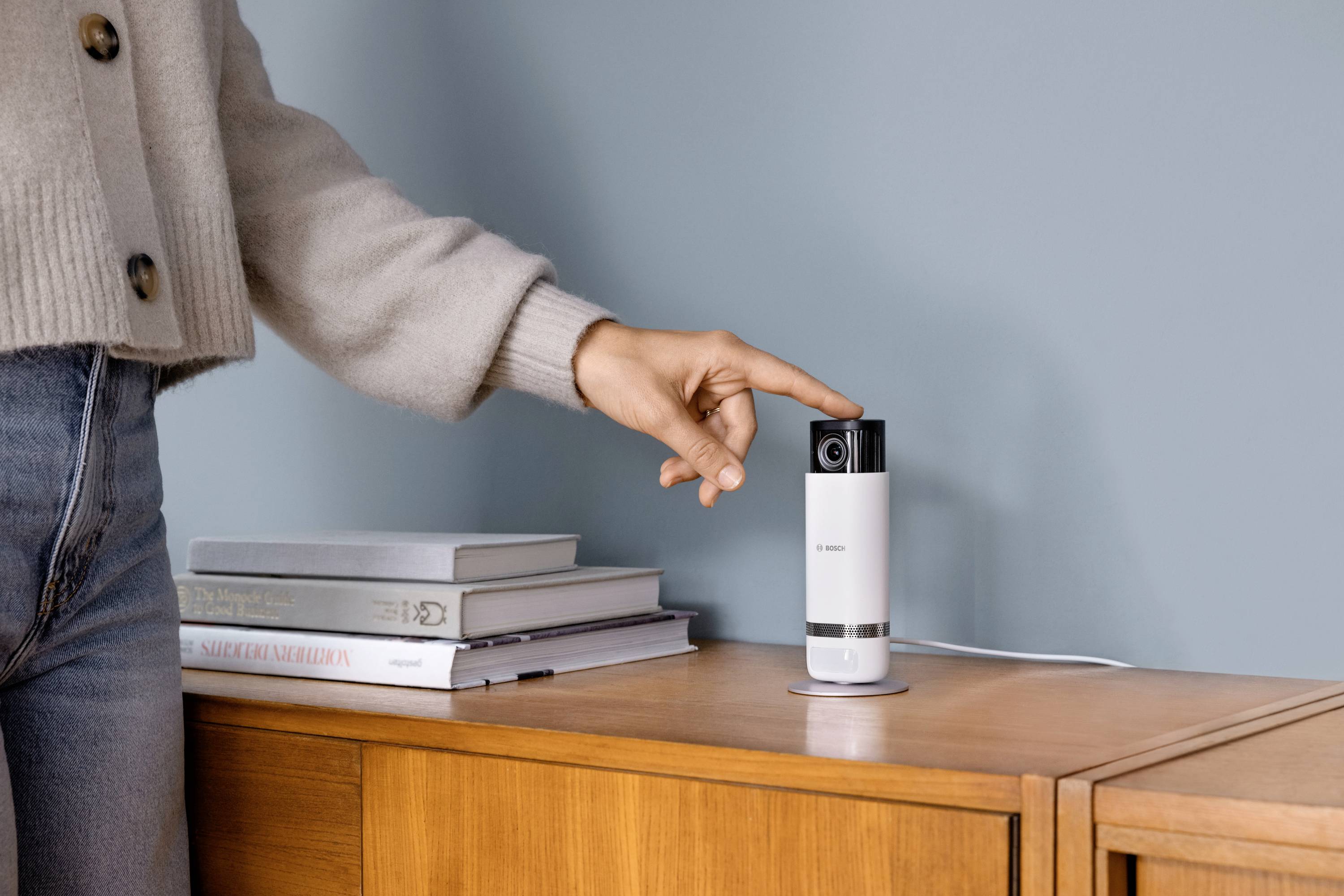 Una persona con abiti casual tocca una piccola telecamera ferma su un mobile in legno con alcuni libri accanto.