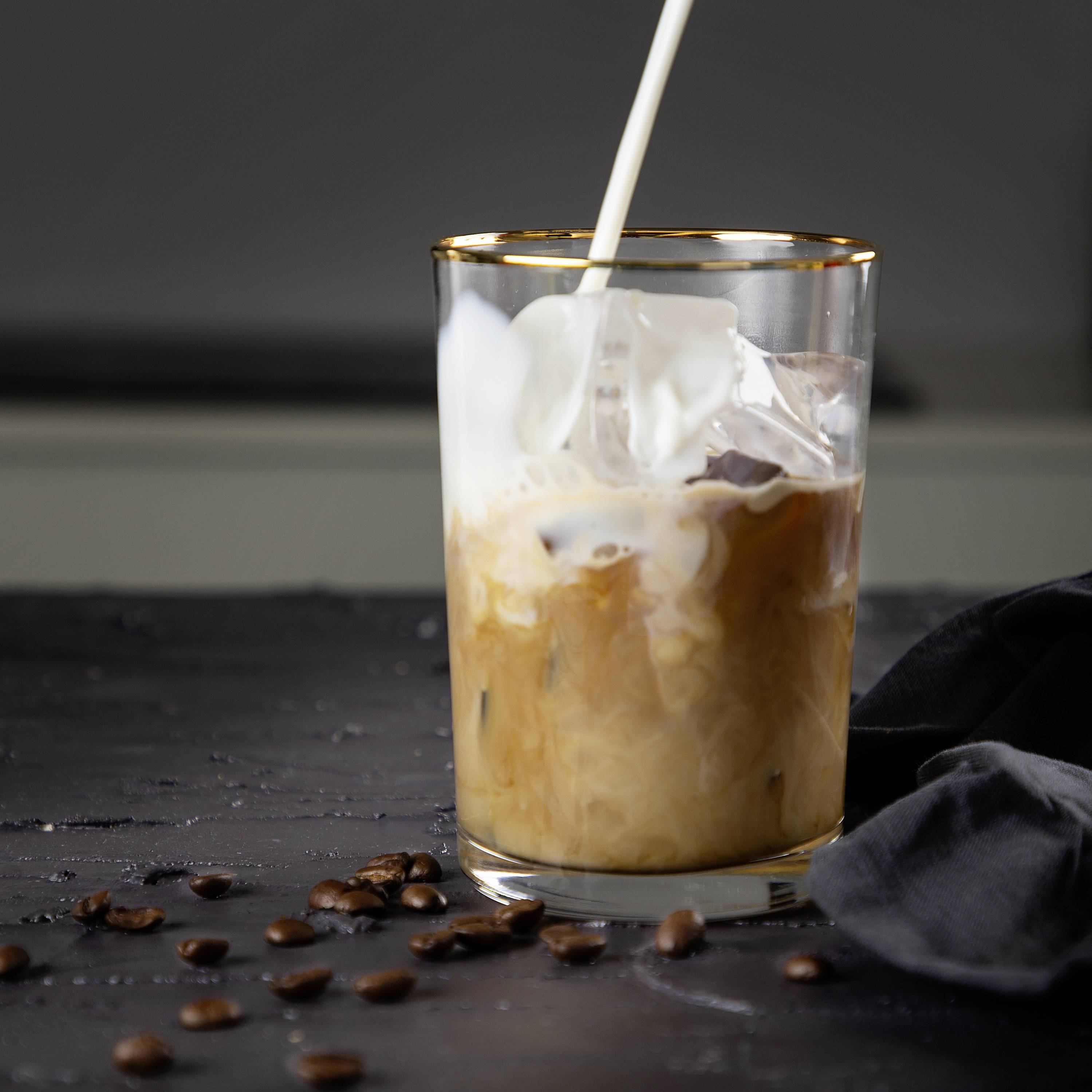 Un bicchiere di caffè freddo con panna e cannuccia è posizionato su un tavolo. Alcuni chicchi di caffè sono sparsi accanto, sullo sfondo sfocato.