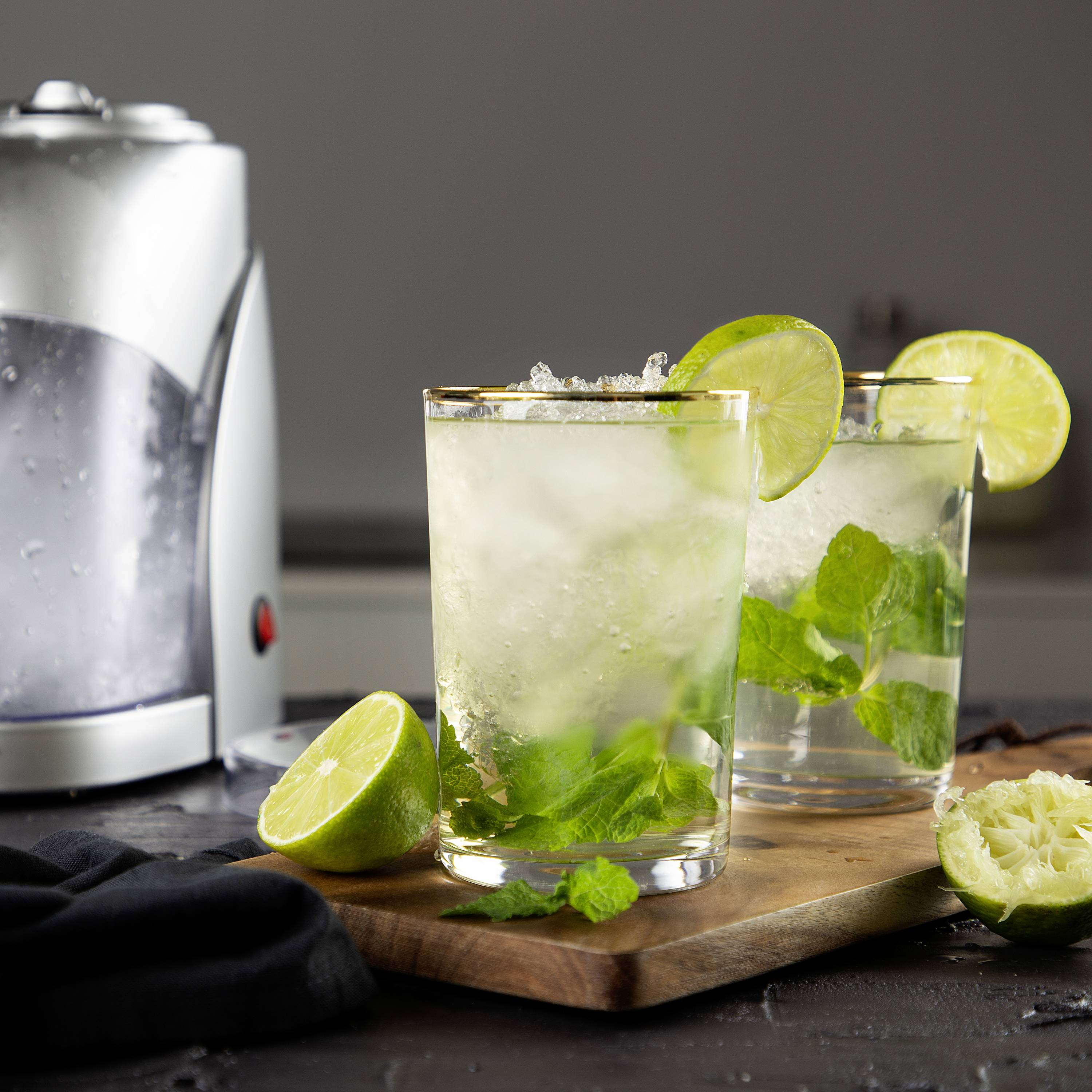 Due bicchieri di cocktail Mojito su un tagliere di legno, guarniti con fette di lime e foglie di menta, accanto a un premilimoni.