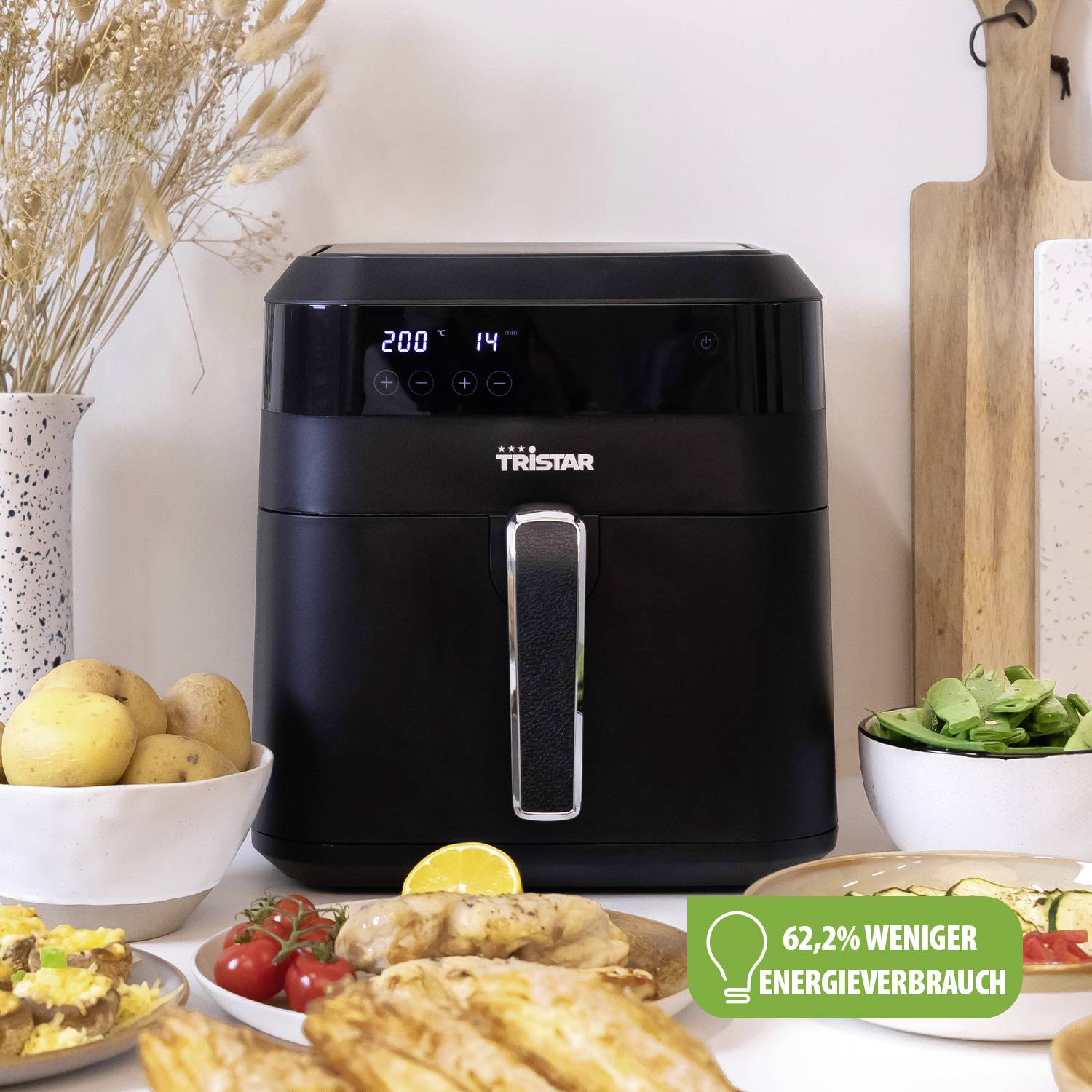 Una friggitrice nera con display digitale è posizionata su un tavolo, circondata da verdure e pane. Un simbolo indica un consumo energetico ridotto del 62,2%.