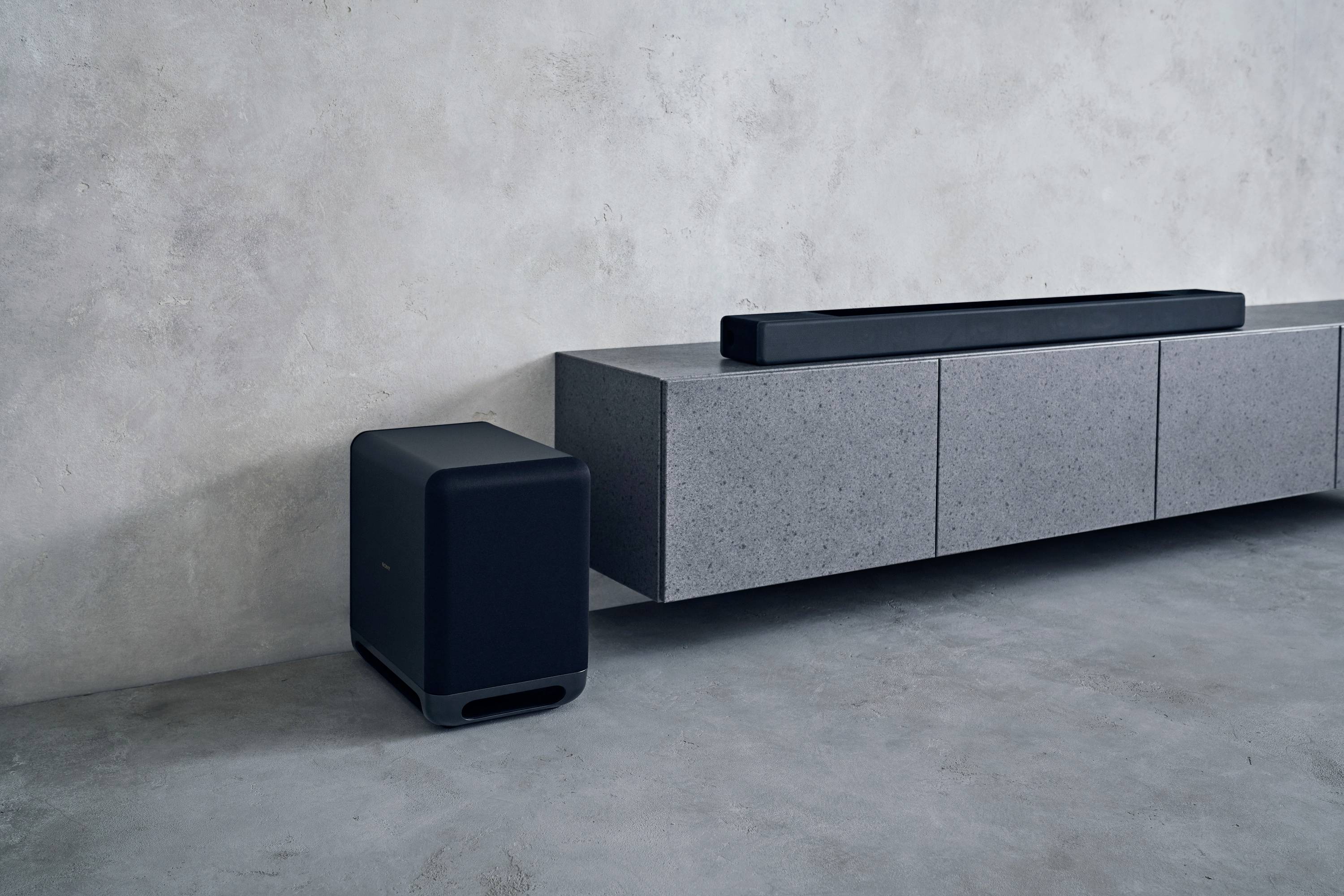 Un soggiorno moderno con un mobile TV grigio appoggiato a una parete. Sopra di esso è posizionata una soundbar nera. Accanto, un subwoofer è poggiato sul pavimento.