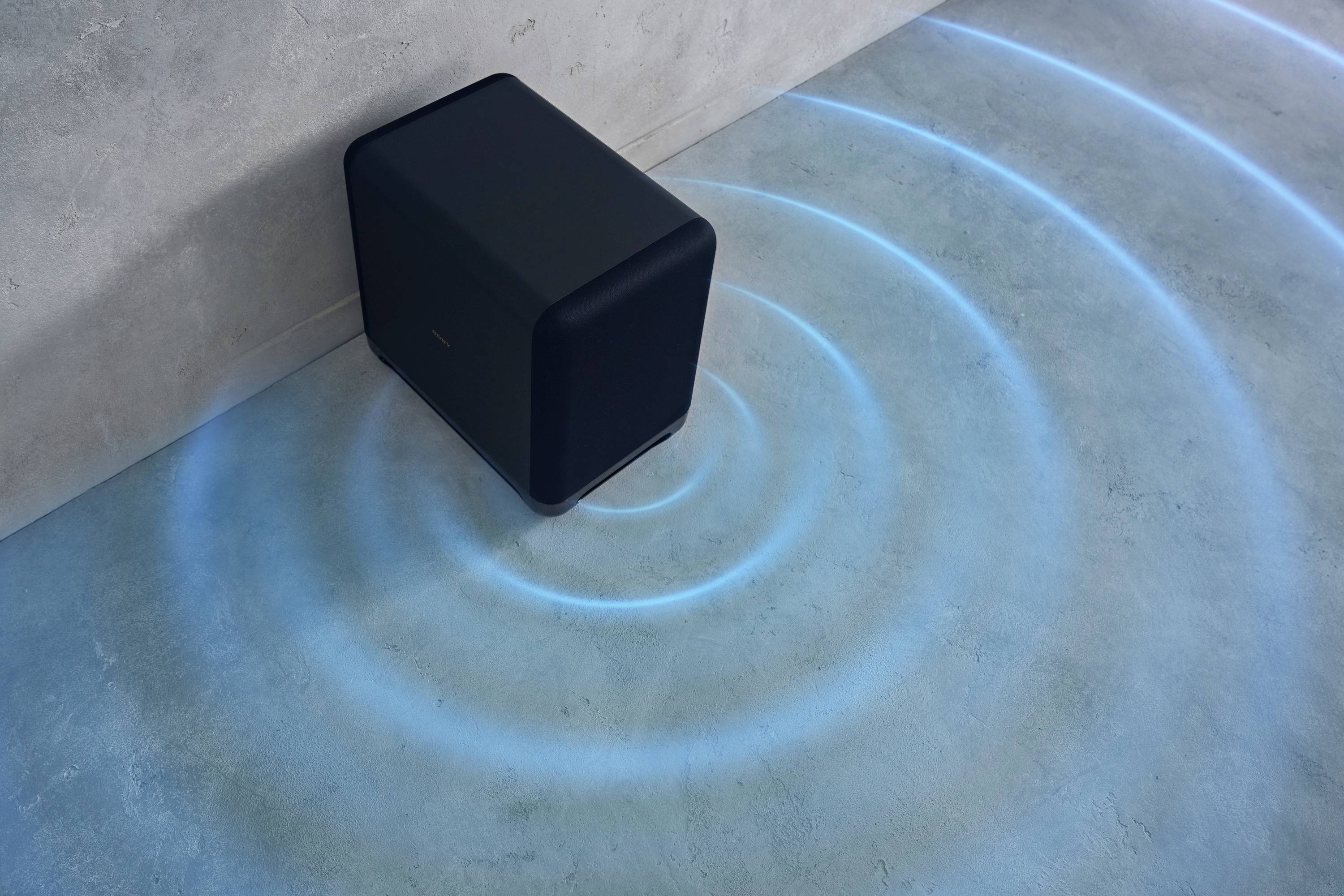 Un altoparlante nero è posizionato nell'angolo di una stanza. Onde sonore blu si propagano concentricamente intorno all'altoparlante sul pavimento.