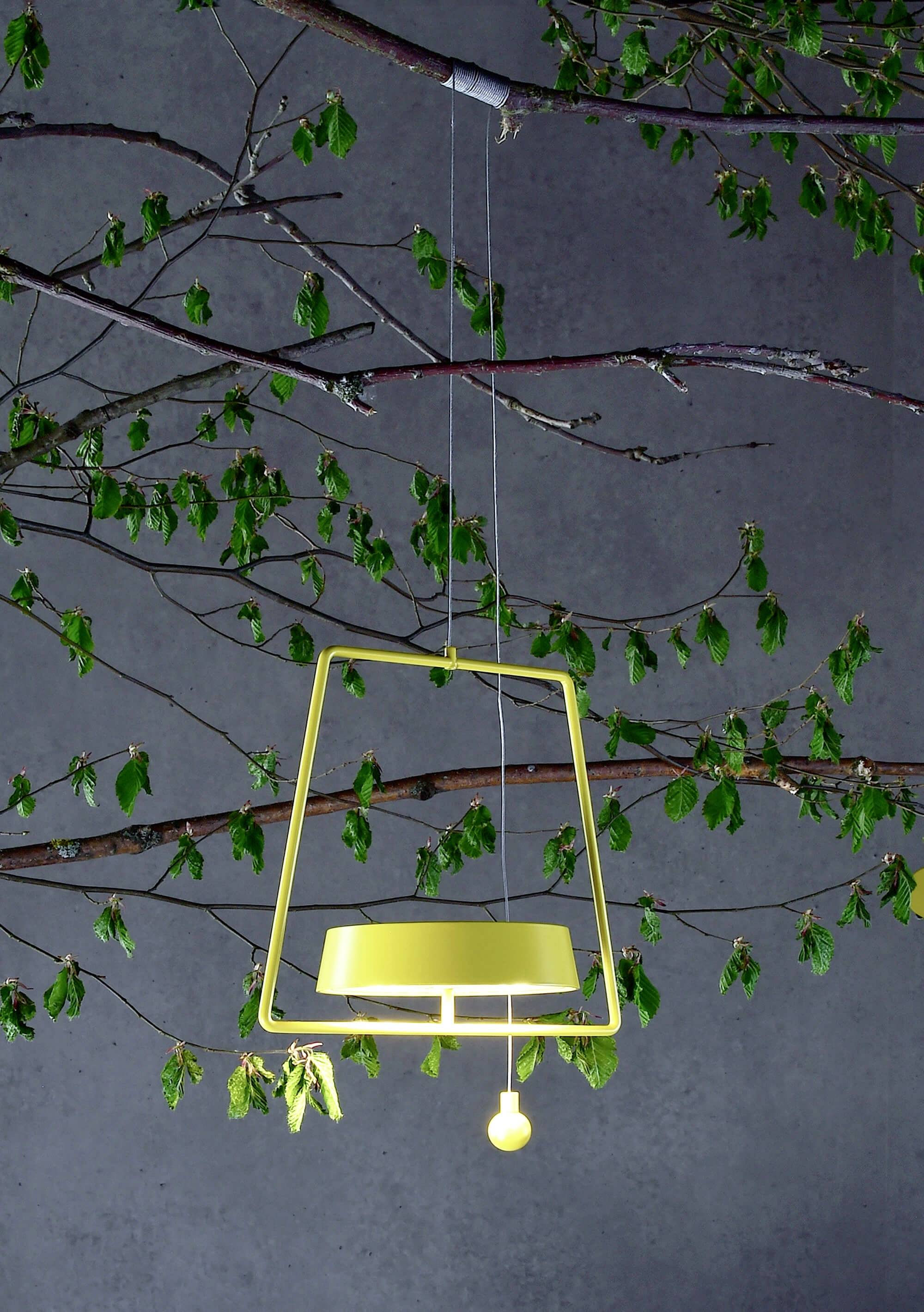 Una lampada a sospensione gialla dalla forma geometrica illumina uno sfondo con rami e foglie verdi.
