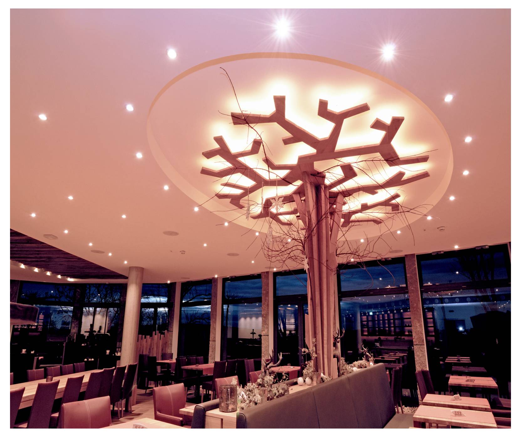 Interno di un moderno ristorante con un lampadario a forma di albero luminoso al centro del soffitto, sopra tavoli da pranzo ordinatamente apparecchiati e sedie.