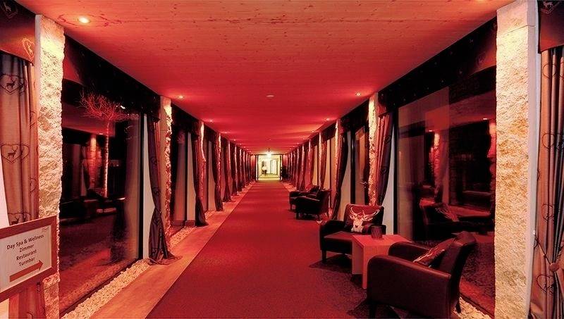 Un lungo corridoio rosso con un divano e poltrone ai lati, decorato con tende e pietre, conduce a una porta illuminata.
